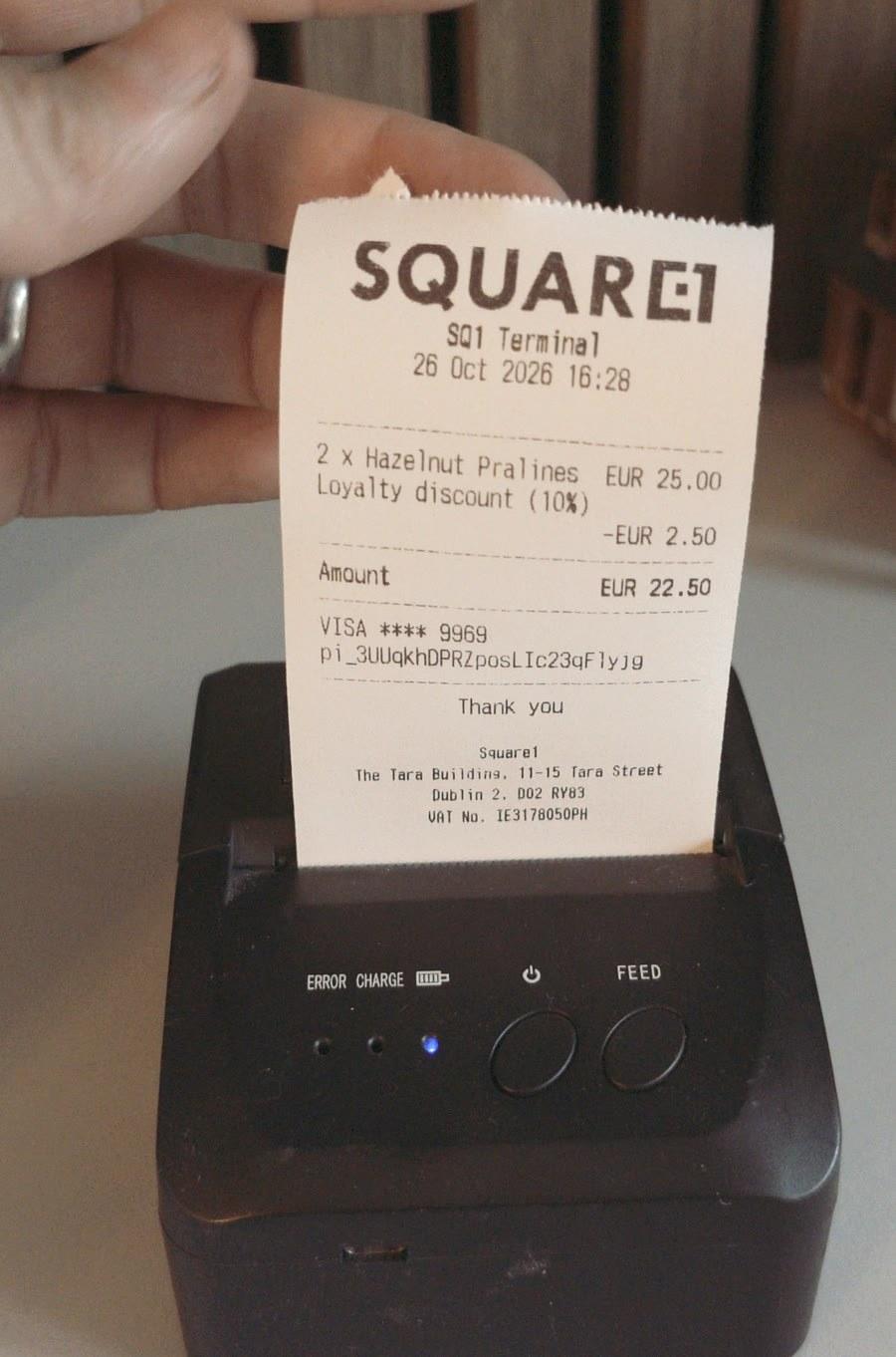 A receipt coming out of a small black Bluetooth thermal printer, with the Square1 logo, 2 x Hazelnut Pralines at EUR 25.00, a 10% loyalty discount of minus EUR 2.50, an amount of EUR 22.50, the card and PaymentIntent ID